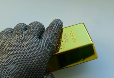 Close-up of a metal gloved hand holding a shiny gold bar indoors.